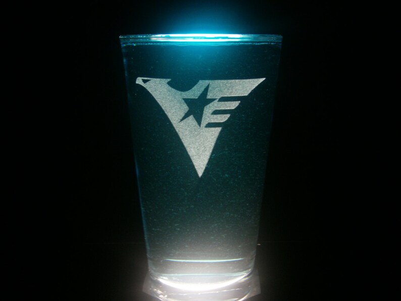 K&ouml;nnte beinhalten: Ein klares Glas mit einem mattierten Design eines Sterns und des Buchstabens "E" an der Seite. Das Glas ist mit einer blauen Fl&uuml;ssigkeit gef&uuml;llt.