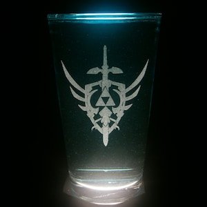 May include: A clear glass with a white etched design of a sword and shield with a triangular symbol. The glass is filled with a blue liquid.