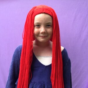 May include: A young person with a red wig and a blue top against a purple background. The wig is long and straight, covering the shoulders. The blue top has a v-neck and long sleeves. The background is a solid purple color.
