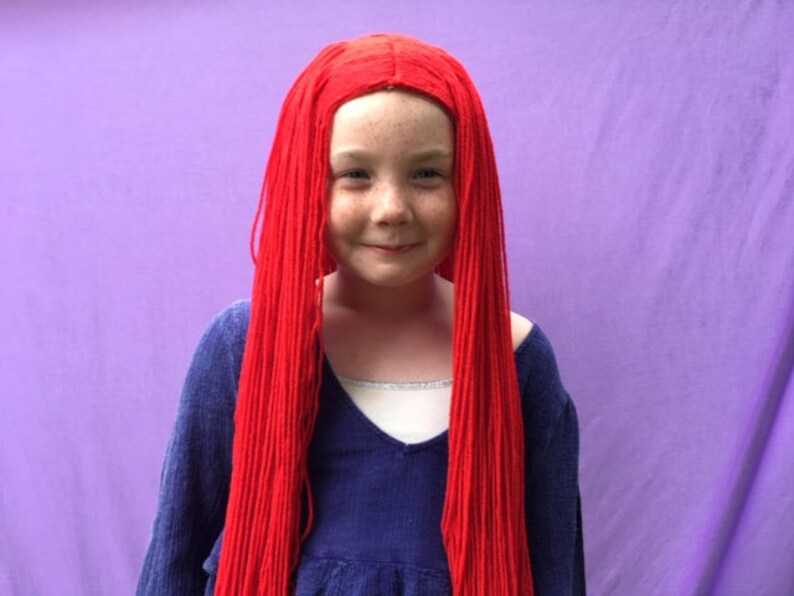 May include: A young person with a red wig and a blue top against a purple background. The wig is long and straight, covering the shoulders. The blue top has a v-neck and long sleeves. The background is a solid purple color.