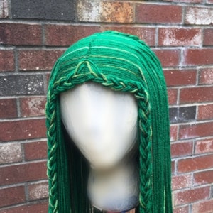 May include: A green wig with long, braided strands. The wig features a braided front and a detailed crown. The color is a vibrant green with subtle variations, creating a textured appearance. The wig is displayed on a mannequin head against a brick wall.