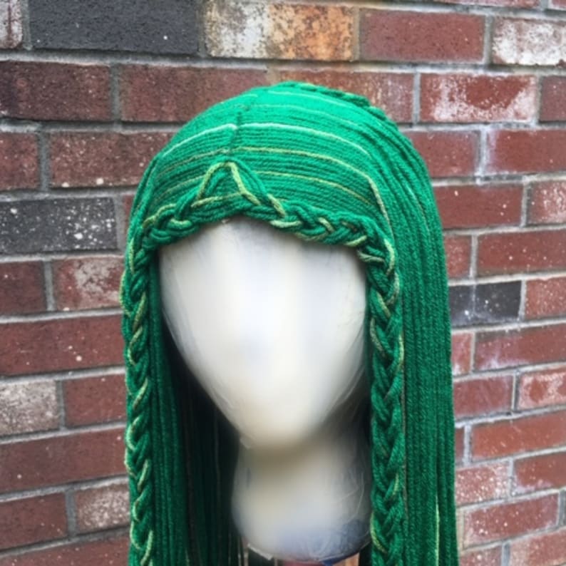 May include: A green wig with long, braided strands. The wig features a braided front and a detailed crown. The color is a vibrant green with subtle variations, creating a textured appearance. The wig is displayed on a mannequin head against a brick wall.