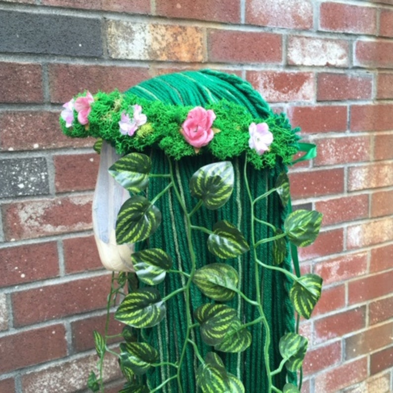 May include: A green yarn wig with a moss and flower crown. The wig has long strands of green yarn with faux green leaves and vines cascading down. Pink and white flowers are placed around the crown.