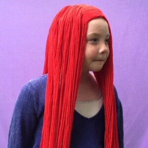 May include: A young person wearing a vibrant red wig with long, straight strands. The wig covers the head and cascades down the shoulders. The person is wearing a blue long-sleeved shirt and a silver necklace. The background is a solid purple.