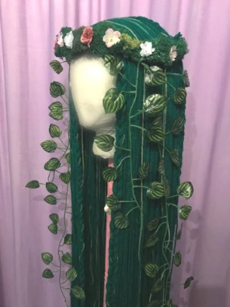 May include: A green wig with long, braided strands, adorned with a floral crown and faux ivy vines. The crown features white, pink, and red flowers. The wig is displayed on a mannequin head against a purple backdrop.