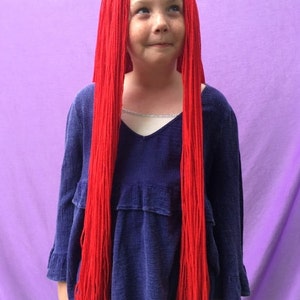 May include: A child wearing a long, bright red wig with a blue long-sleeved top. The wig is made of long, straight strands. The top has a V-neck and ruffled details. The background is a solid purple.