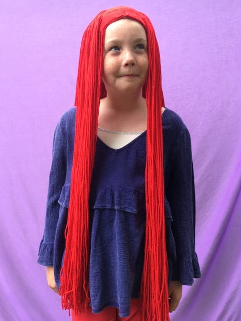 May include: A child wearing a long, bright red wig with a blue long-sleeved top. The wig is made of long, straight strands. The top has a V-neck and ruffled details. The background is a solid purple.