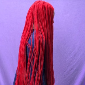 May include: A person is seen from the side, their face obscured by long, vibrant red strands of hair. The hair cascades down, contrasting with a denim jacket. The background is a solid, soft purple.