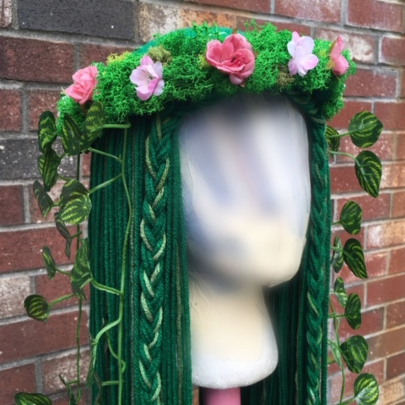 May include: A green wig with long, braided strands, adorned with a moss-covered crown featuring pink flowers and trailing green vines. The wig is displayed on a white mannequin head against a brick wall.