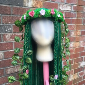 May include: A green wig with long, braided strands, adorned with a floral crown of pink and white flowers and faux ivy vines. The wig is displayed on a mannequin head against a brick wall background. The wig is a costume piece.