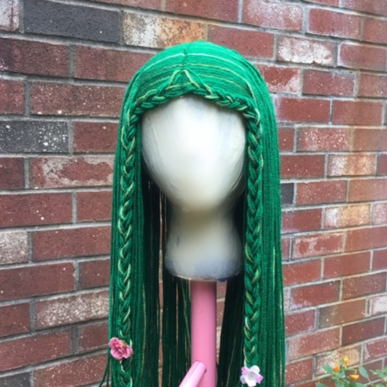 May include: A green wig with long, braided sections and a central part. The wig is displayed on a mannequin head. The braids are accented with lighter green and white strands. Pink flowers are attached to the braids. The background is a brick wall.