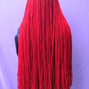 May include: A vibrant red yarn creation, possibly a costume or art piece, cascading down. The strands are long and densely packed, creating a textured, eye-catching effect. The background is a soft purple, providing a contrasting backdrop for the bold red.
