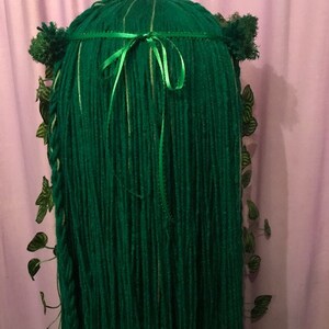 May include: A green costume wig with long, flowing strands. The wig is adorned with a green ribbon, small green pom-poms, and decorative green leaves. The wig is designed to resemble a forest or nature-themed character.