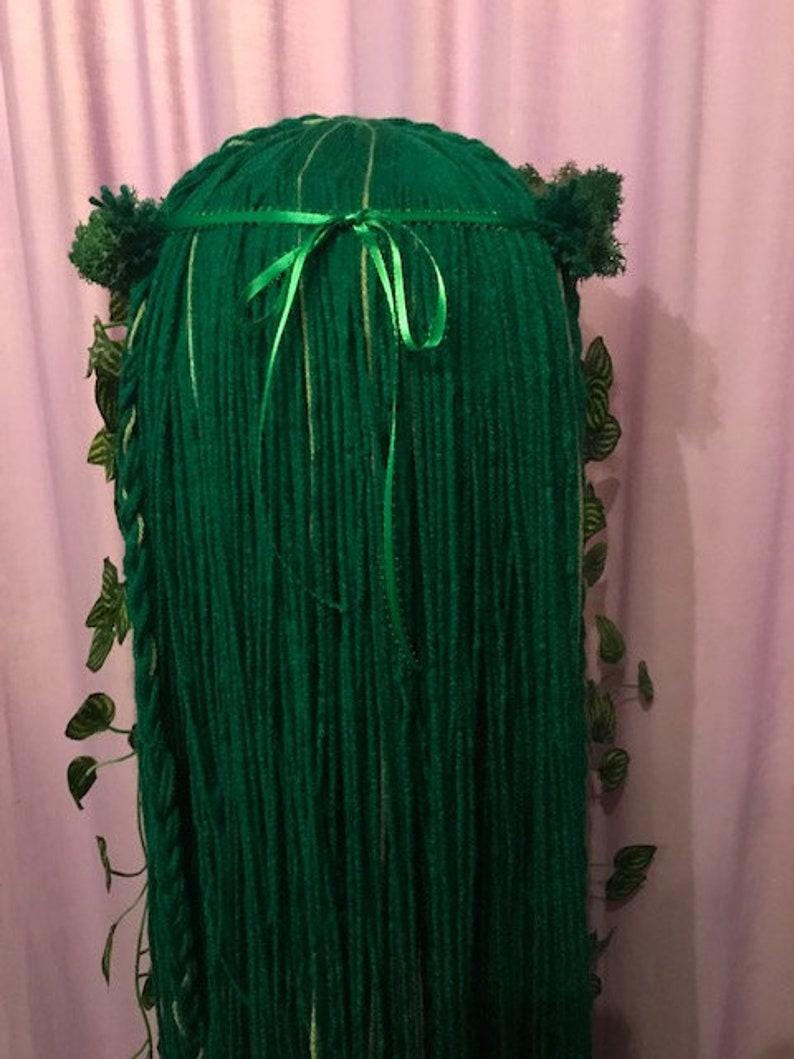 May include: A green costume wig with long, flowing strands. The wig is adorned with a green ribbon, small green pom-poms, and decorative green leaves. The wig is designed to resemble a forest or nature-themed character.