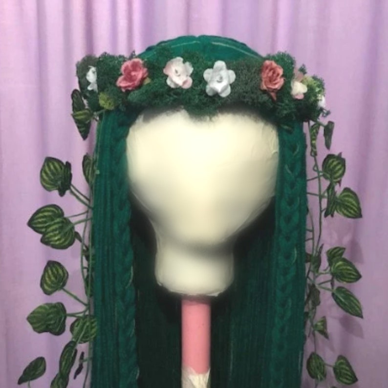 May include: A green wig with long, braided hair, adorned with a moss-covered crown featuring pink and white flowers. Green leaves and vines cascade down the sides, creating a whimsical, nature-inspired look. The wig is displayed on a white mannequin head.