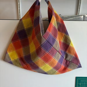May include: A hobo-style bag featuring a multicoloured plaid pattern in shades of orange, yellow, purple, and green. The fabric is accented with white stitched grid lines throughout. The bag has a single handle and a light-coloured interior lining.