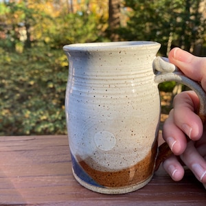 May include: Handmade ceramic mug with a speckled white and brown glaze. The mug features a curved handle and a design with blue and brown accents. The mug is held by a hand.