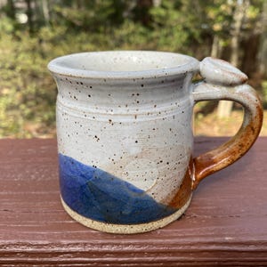 May include: Handmade ceramic mug with a speckled white glaze, a blue and brown color block design, and a unique handle. The mug is approximately 4 inches tall and 3.5 inches in diameter, perfect for coffee or tea.