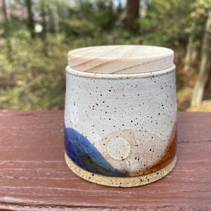 May include: A ceramic jar with a wooden lid. The jar is cylindrical with a speckled white glaze, accented with blue, brown, and tan color blocks. The lid is made of light-colored wood. The jar sits on a brown wooden surface.