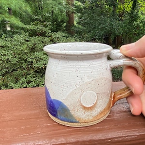 May include: Handmade ceramic mug with a speckled white glaze, featuring blue and tan abstract designs. The mug has a rounded shape, a sturdy handle, and a small, circular accent. The mug is sitting on a brown surface.