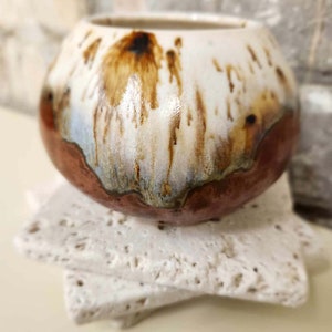 May include: A white ceramic pot with brown and orange glaze, sitting on a white ceramic base. The pot has a rounded shape and a wide opening.