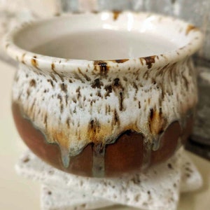 May include: A small, brown ceramic pot with a white glaze and brown speckles. The pot has a rounded shape and a wide opening. It is sitting on a white, star-shaped base.