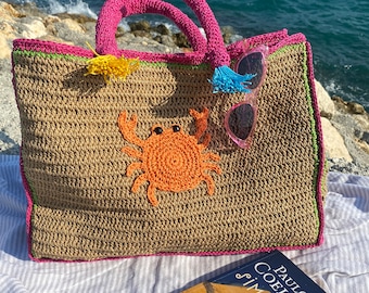 May include: A woven beach tote bag with a crab applique, pink handles, and a book. The bag has a brown body with pink and green trim. Pink sunglasses and a book titled "Paulo Coelho Simyaci" are next to the bag.