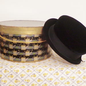 May include: A vintage black top hat rests beside a round hat box. The hat box has a gold-painted rim and a patterned design in black, green, and gold. The box sits on a patterned tablecloth.
