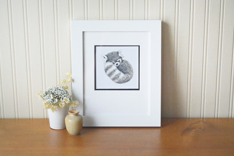 Red Panda Art print. This photo shows and example of how you could frame the art. It's in a white frame, sitting on a wooden table with small vases to the left of the frame. Red panda curled up sleeping. Fluffy tail is in front.