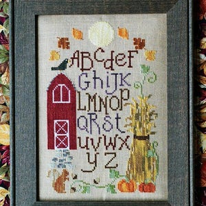 Peut inclure: Un sampler de point de croix représentant l'alphabet dans une scène rustique d'automne. Les lettres sont brodées en violet et bleu, avec une grange rouge, un épouvantail et des citrouilles en arrière-plan.