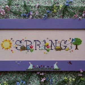 May include: A purple framed cross-stitch with the word "SPRING" in colorful embroidery. The design includes a sun, flowers, a bunny, a bird, and a kite.