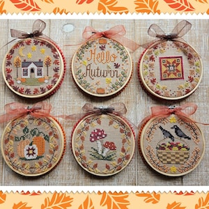May include: Six embroidered hoops with autumnal designs. Each hoop features a different image, including a house, the text "Hello Autumn", a quilt pattern, pumpkins, mushrooms, and a basket with birds. The hoops are decorated with fall leaves and tied with ribbons.