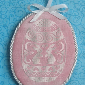 May include: A pink oval-shaped ornament with a white embroidered design. The ornament is trimmed with a white rope-like border and has a white ribbon attached.