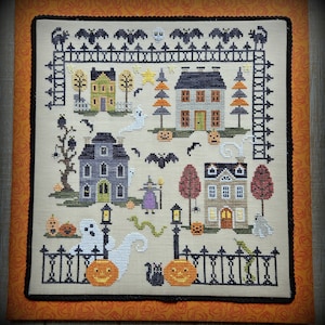 Puede incluir: Un patrón de punto de cruz que representa un pueblo de Halloween con casas, árboles, calabazas, murciélagos y un fantasma. El patrón está enmarcado en un marco negro y naranja.