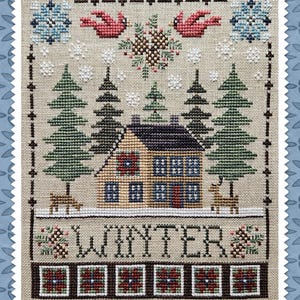 May include: A cross-stitch embroidery on beige fabric featuring a winter scene with a house, pine trees, two deer, red birds, and snowflakes. The word WINTER is stitched in black letters above a row of five decorative red and green squares.