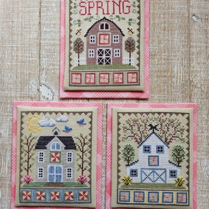 May include: Three cross-stitch embroidery designs featuring a red barn, a white barn, and a red barn with the word "SPRING" above it. Each design is framed with a pink and white gingham border.