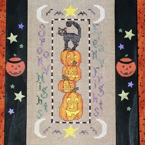 May include: Framed Halloween cross-stitch featuring a black cat atop stacked jack-o'-lanterns. The artwork includes stars, moons, and text, all within a black frame. The background is tan, with orange and black accents.