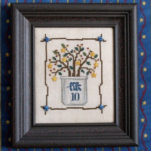 Op de afbeelding: Een ingelijst kruissteekontwerp met een plant in een pot met gele bloemen en groene bladeren. De pot is blauw en wit met het nummer "10" erop.