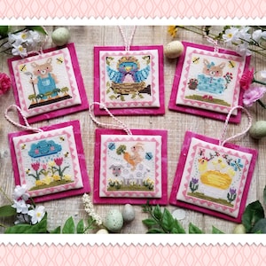 May include: A collection of six square cross-stitch ornaments with Easter themes. Each features a different design: a bunny, a bird, a sheep, a chick, a cloud with rain, and a bunny. The ornaments are set on pink fabric with white string hangers.