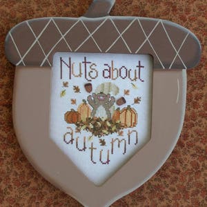 Puede incluir: Un marco de madera marrón en forma de bellota con un fondo blanco y un diseño de punto de cruz. El diseño presenta una ardilla sosteniendo una nuez, calabazas y hojas. El texto "Nuts about autumn" está bordado en hilo marrón.