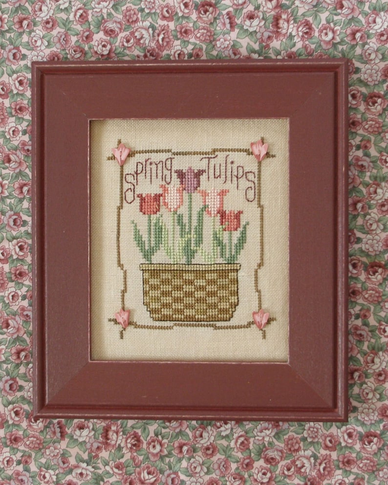 May include: A framed cross-stitch embroidery depicting a pot of pink and red tulips with green leaves. The text "Spring Tulips" is stitched above the pot.