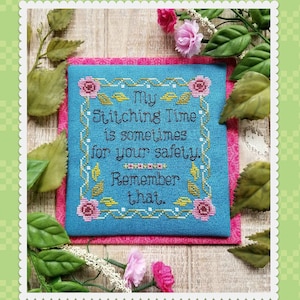 以下が含まれることがあります： ピンクの花と金の枠が付いた青いクロスステッチのサンプル。テキストには「私の縫い時間は、あなたの安全のためでもあります。覚えておいてください。」と書いてあります。