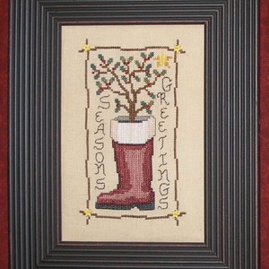May include: A cross-stitch design featuring a red boot with a green holly plant growing out of it. The boot is filled with white stuffing and the plant has red berries. The design is framed in a black frame and the text "Seasons Greetings" is stitched on the right side of the design.