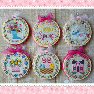 May include: Six round embroidery hoops with spring-themed designs. The hoops are decorated with pink and white ribbon. The designs include a bouquet of tulips in a blue boot, a basket of Easter eggs, a bunny rabbit, a house, a watering can with flowers, and the words "Hello Spring".