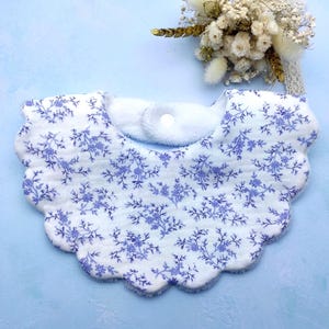 Könnte beinhalten: Ein weißes Baby-Lätzchen mit blauem Blumenmuster und gewelltem Rand. Das Lätzchen hat eine weiche, saugfähige Textur und einen Druckknopfverschluss. Ein kleiner Strauß Trockenblumen befindet sich im Hintergrund.