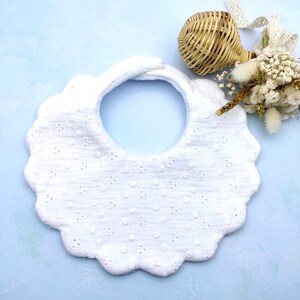 Könnte beinhalten: Weißes Baby-Lätzchen mit gewelltem Rand und Lochmuster. Das Lätzchen hat eine runde Halsöffnung und einen Klettverschluss. Ein kleiner Weidenkorb und Trockenblumen befinden sich im Hintergrund, auf einer hellblauen Oberfläche.