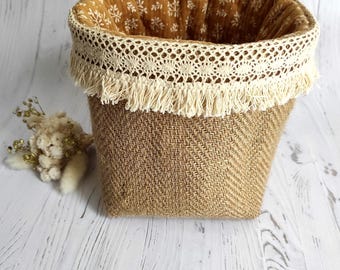 May include: A cylindrical storage basket featuring a textured tan herringbone weave exterior. The top edge is trimmed with cream-colored lace and fringe. The interior is lined with a mustard yellow fabric displaying a white botanical pattern.