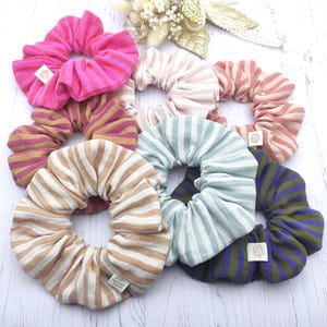Peut inclure: Plusieurs chouchous rayés de différentes couleurs, dont rose, beige, bleu et vert. Chaque chouchou est orné d'une étiquette en tissu. Un petit bouquet de fleurs séchées et un nœud doré sont en arrière-plan.