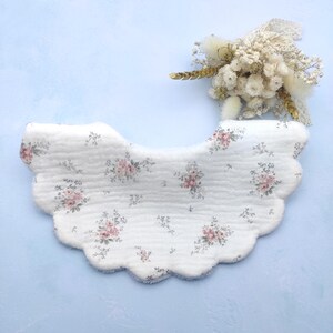 Könnte beinhalten: Ein weißes Baby-Lätzchen mit gewelltem Rand, verziert mit einem zarten Blumenmuster in Rosa und Grün. Das Lätzchen ist aus weichem Stoff gefertigt und vor einem hellblauen Hintergrund platziert, mit einem kleinen Strauß Trockenblumen.