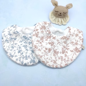 Puede incluir: Dos baberos para bebé con estampados florales en azul y marrón, junto con un juguete de conejo de punto. Los baberos tienen un fondo blanco con delicados diseños de hojas y flores. El conejo es beige con los ojos cerrados y un cuello con volantes.